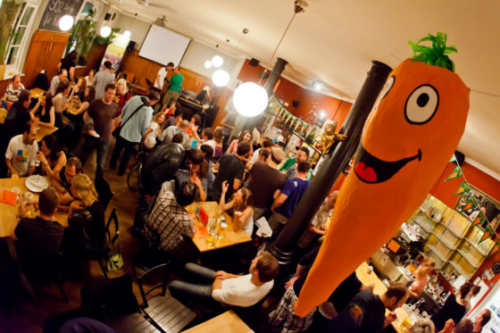 Menschen feiern in einer Kneipe, im Vordergrund eine überdimensionale Möhre