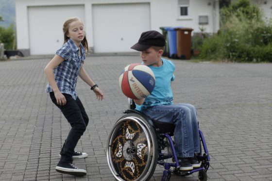 Zwei Kinder spielen mit einem Ball. Das Mädchen steht, der Junge mit dem Ball sitzt im Rollstuhl.