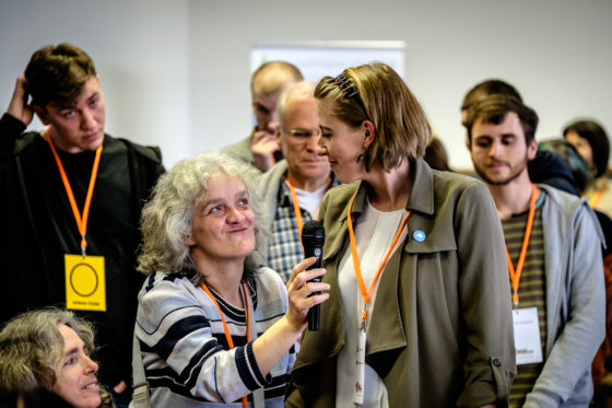 Das Foto zeigt Teilnehmende des openTransfer CAMP #Inklusion. Im Vordergrund hält eine weiblich gelesene Person, einer anderen weiblich gelesenen Teilnehmerin ein Mikrofon hin. Beide lächeln. Sie tragen orangene Lanyards um den Hals.