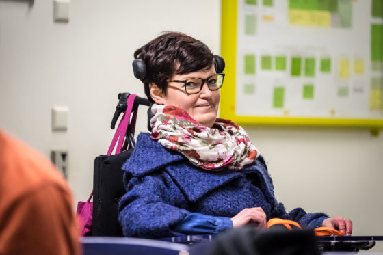 Das Foto zeigt Anastasia Umrik, Teilnehmerin des openTransfer CAMPs #Inklusion. Sie trägt kurze braune Haare, eine Brille, bunten Schal und marineblaue Jacke. Sie sitzt in einem Rollstuhl und lächelt in die Kamera. Im Hintergrund ist ein Whiteboard mit Post-Its darauf zu sehen.