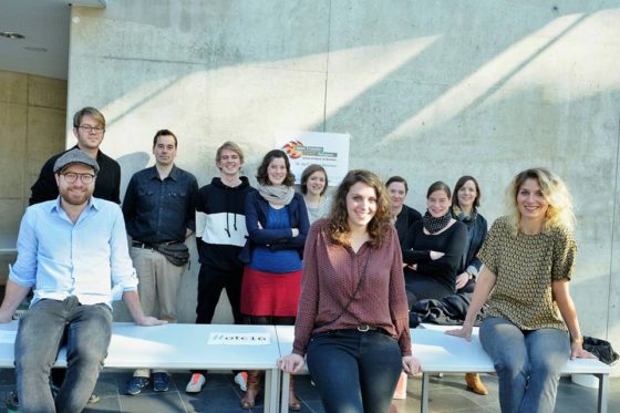 Ein Dutzend junger Menschen hat sich zum Gruppenfoto vor einer Betonwand aufgstellt.