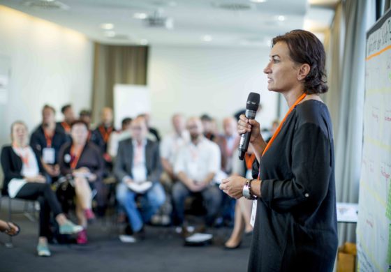 Eine Frau mit Mikrofon steht vor der sitzenden Zuhörerschaft.