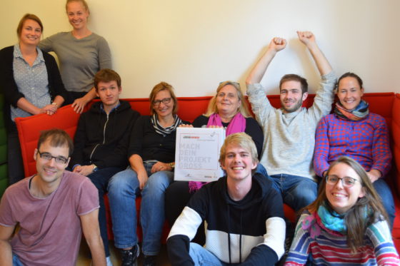 Eine Gruppe von Menschen sitzt auf einem Sofa. in der Hand hält eine Person einen Ordner auf dem draufsteht Mach Dein Projekt. Alle schauen in die Kamera.