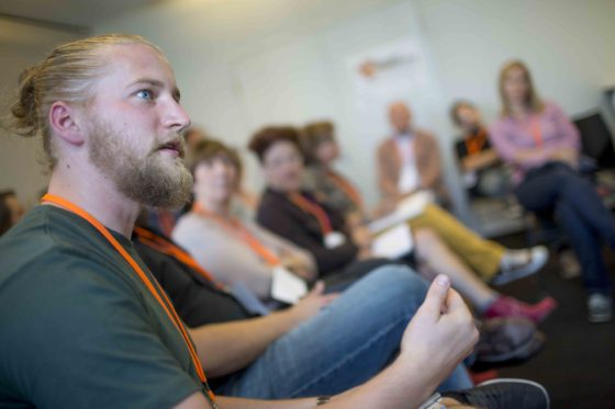 Ein blonder junger Mann erklärt den um ihn Herumsitzenden etwas.