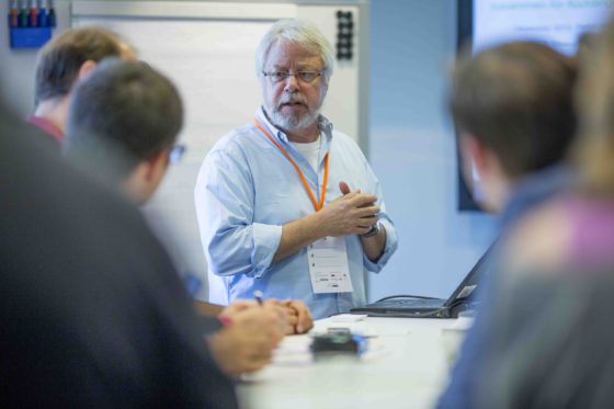 Ein Mann mit weißen Haaren steht vor einer Gruppe Zuhörer und erzählt.