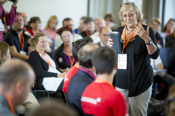 Eine ältere Frau mit Brille spricht vor einem sitzenden Publikum.