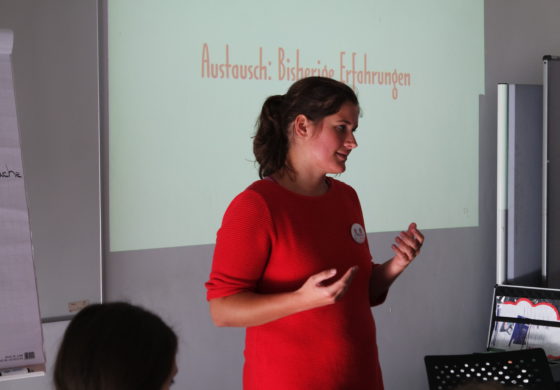 Eine Frau in rotem Kleid erklärt etwas vor einer Leinwand.