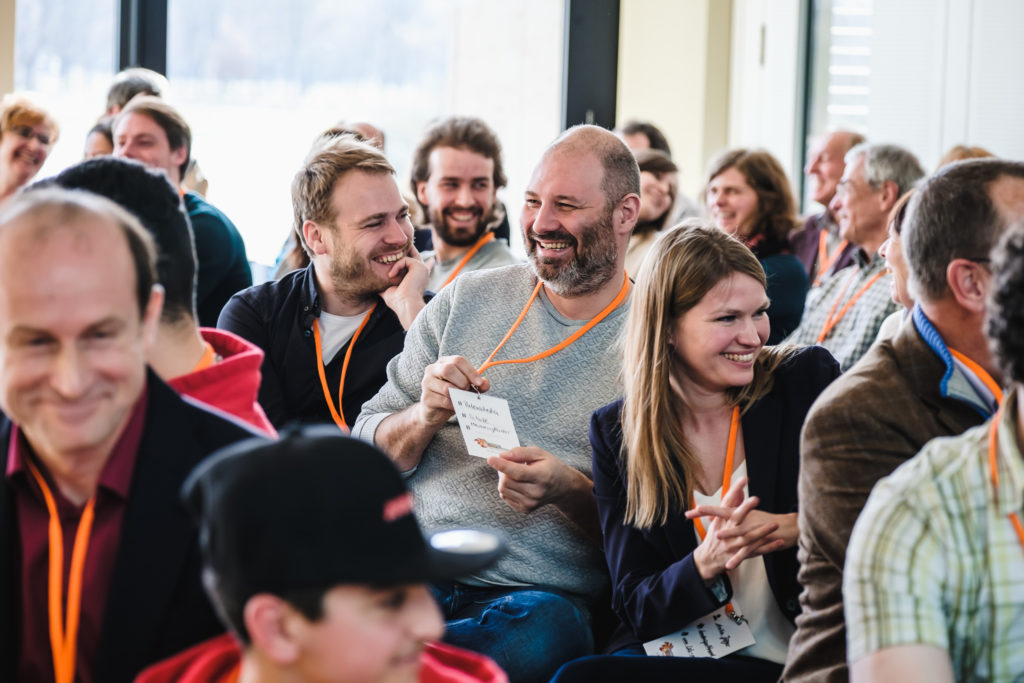 Eine Gruppe von Menschen sitzt im Publikum einer Veranstaltung. Die Personen lachen und schauen nach links.