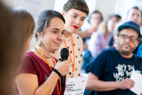 Ein Frau hält ein Mikrofon und ist von mehreren Menschen umgeben.