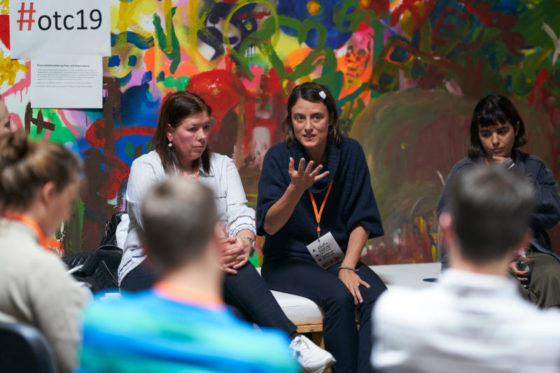 Eine Frau sitzt vor einer Wand mit Grafittis und erklärte einem Stuhlkreis etwas.