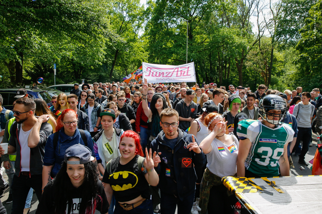 Das Foto zeigt eine Menschenmenge beim CSD. Die Menschen tragen Plakate vor sich her, sind verkleidet und tragen Regenbogenflaggen auf ihrer Kleidung.