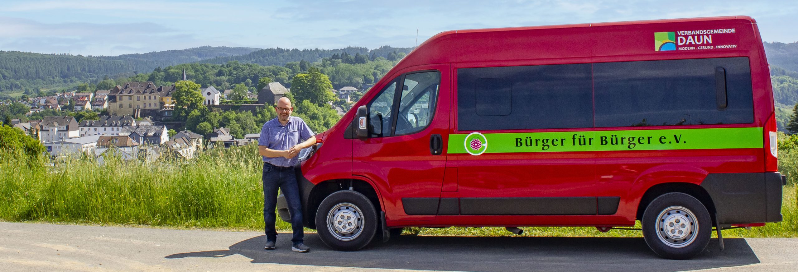 Auf dem Foto steht ein Mann neben einem roten Bus. Auf dem Bus steht die Aufschrift "Bürger für Bürger e.V.". Im Hintergrund ist die Landschaft der Vulkaneifel zu sehen.