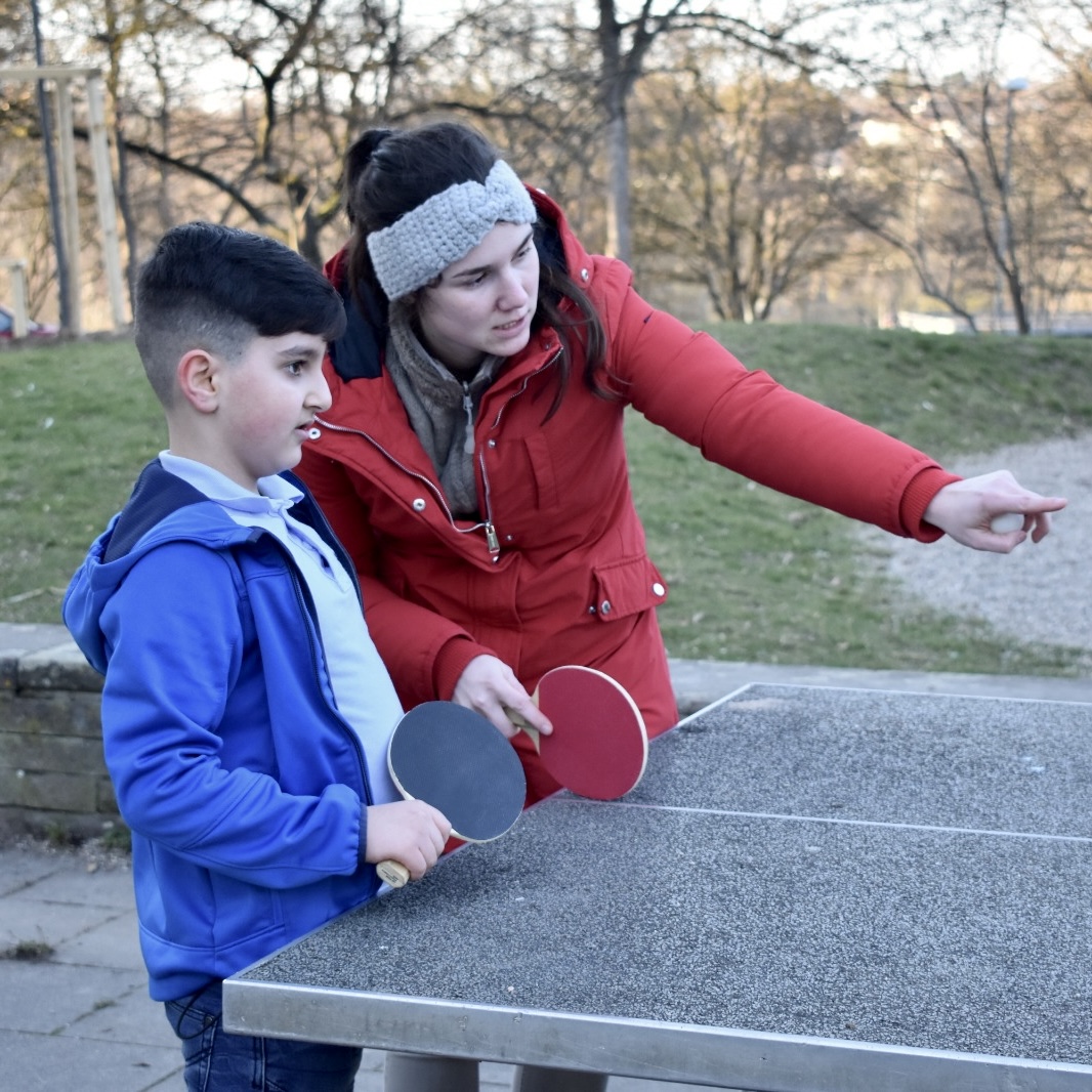 Bild II: Auf dem Foto ist ein kleiner Junge und eine erwachsene Frau zu sehen. Beide stehen nebeneinander an einer Tischtennisplatte und haben einen Tischtennisschläger in der Hand. Die Frau zeigt mit ihrem Arm auf etwas und scheint dem Jungen etwas zu erklären.