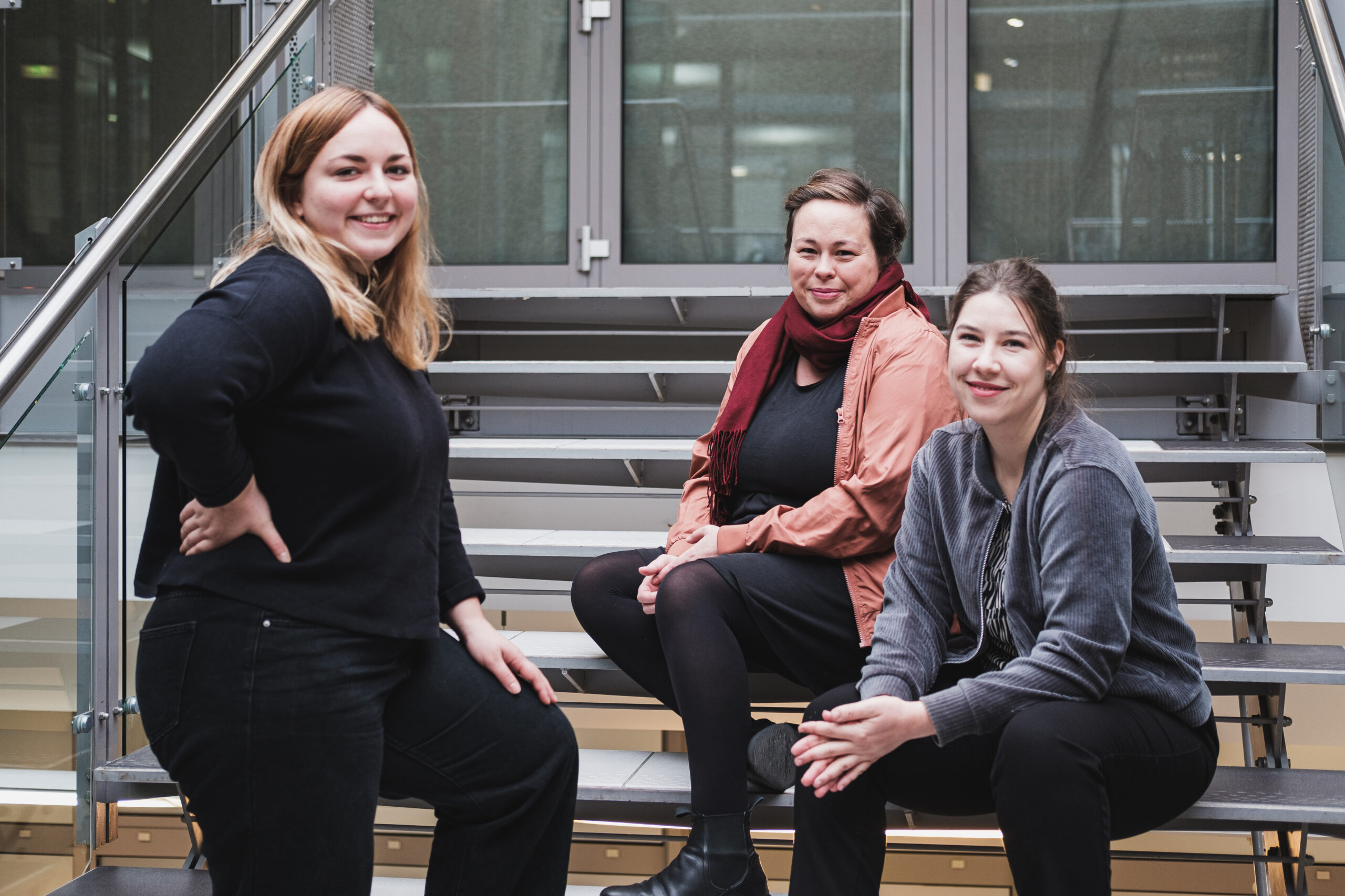 Teamfoto openTransfer Zusammenhalt: Rechts im Bild sitzen zwei weiblich gelesene Personen auf einer Treppe, links im Bild ist eine dritte weiblich gelesene Person zu sehen. Alle drei schauen in die Kamera und lächeln.