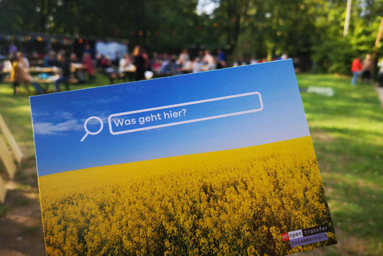 Bild einer Postkarte, auf der ein gelbes Rapsfeld und das Logo von openTransfer Zusammenhalt zu sehen ist. Dahinter verschwommen Menschen beim Essen auf einem Festival