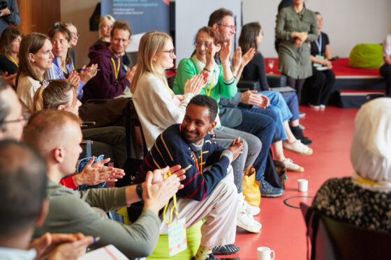 Mehrere Personen sitzen in einem Halbkreis in einem Raum mit rotem Boden und klatschen. Einige tragen gelbe Bänder um den Hals. Im Vordergrund ist der Rücken einer Person mit weißem Kopftuch zu sehen. Im Hintergrund stehen weitere Personen.