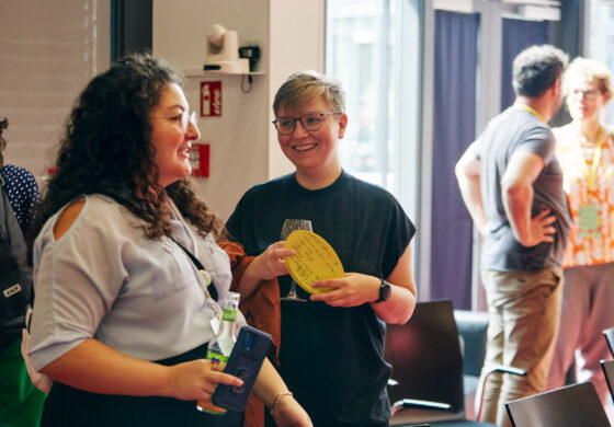 Zwei Personen stehen in einem hellen Raum und unterhalten sich. Eine Person hält eine gelbe Karte mit handgeschriebenem Text und ein Weinglas, die andere eine Flasche und ein Smartphone. Im Hintergrund sind weitere Personen im Gespräch sowie Stühle und große Fenster zu sehen.