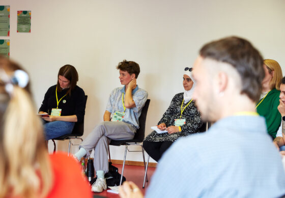 Das Foto zeigt eine Gruppe von Personen, die in einem Halbkreis auf schwarzen Stühlen sitzen. Sie tragen gelbe Lanyards um den Hals. Einige machen sich Notizen.