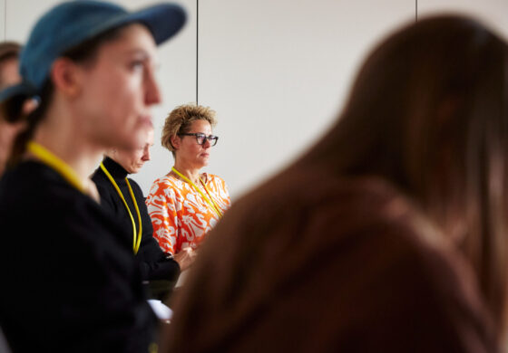 Mehrere Personen sitzen in einem Raum und schauen in eine Richtung. Eine weiblich gelesene Person in einem orange-weiß gemusterten Oberteil mit gelbem Band um den Hals sitzt in der Mitte des Bildes. Im Vordergrund sind weitere Personen unscharf zu sehen.