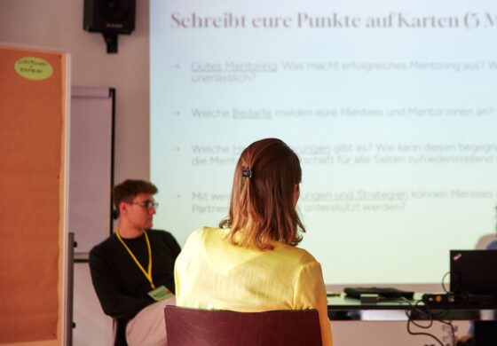 Das Foto zeigt zwei Personen, die von hinten, bzw. seitlich abfotografiert wurden. Beide sitzen auf Stühlen und schauen in Richtung einer PPP, die an die Wand projiziert wurde. Am rechten Rand des Fotos ist ein Tisch mit einem aufgeklapptem Laptop zu sehen, sowie eine Person, die dahinter sitzt.