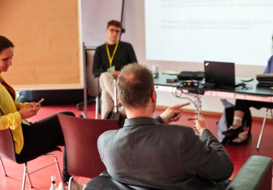 Das Foto zeigt mehrere Personen, die auf Stühlen einander gegenüber sitzen. Eine davon schaut auf ihr Hände, eine andere trägt ein gelbes Lanyard und hört einer dritten, sprechenden Person, zu. An der Wand ist eine Power Point Präsentation zu sehen.