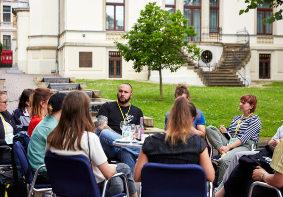 Das Foto zeigt eine Gruppe von Personen, die um einen Tisch herumsitzen und ins Gespräch vertieft sind. Alle tragen gelbe Lanyards um den Hals. Im Hintergrund sieht man eine große, parkähnliche Grünfläche und ein hochherrschaftliches Haus.