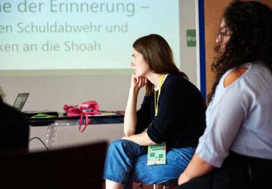 Eine weiblich gelesene Person sitzt in einem Seminarraum und hört konzentriert zu, im Hintergrund ist eine Präsentationsfolie mit Text zur Erinnerungskultur zu sehen. Auf dem Tisch liegen Laptops und Taschen, um den Hals trägt sie ein gelbes Lanyard mit Namensschild.