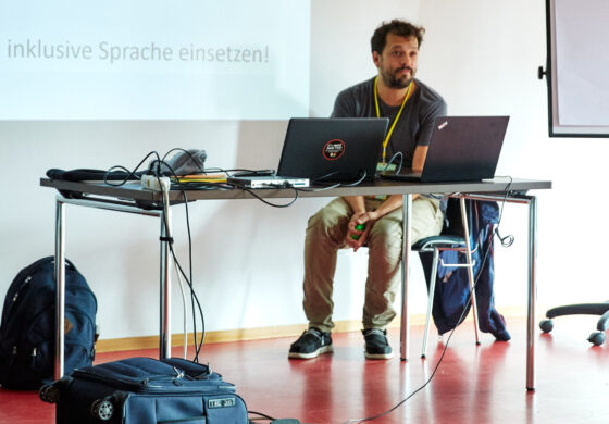 Das Foto zeigt im Vordergrund eine Reihe von Kinderbüchern, die verteilt auf dem Boden liegen. Dahinter sitzt eine männlich gelesene Person an einem Tisch, auf dem zwei geöffnete Laptops stehen. Im Hintergrund ist eine Power Point Präsentation an die Wand projiziert.