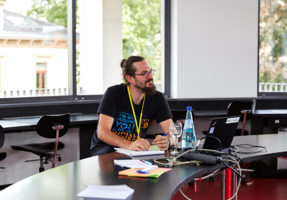 Das Foto zeigt eine männlich gelesene Person, die an einem langen, gebogenem Tisch sitzt. Sie trägt ein gelbes Lanyard um den Hals, trägt eine Brille und hat einen Stift in der Hand. Vor ihr stehen ein Laptop sowie ein Glas und eine Flasche Wasser. Im Hintergrund sieht man helle Glasfronten, sowie ein anderes Haus und Bäume.