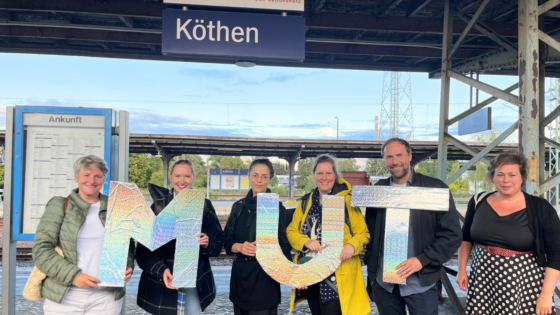 Eine Gruppe von sechs Personen steht auf einem Bahnsteig unter dem Stationsschild „Köthen“. Sie halten gemeinsam große, schillernde Buchstaben, die das Wort „MUT“ bilden. Im Hintergrund sind Gleise, Bahnsteigüberdachungen und der Himmel mit leichten Wolken zu sehen.