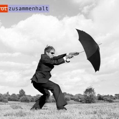 Die schwarz-weiß Fotografie zeigt eine männlich gelesene Person im Anzug, die einen Regenschirm in den ausgestreckten Armen hält und gegen den Wind kämpft. Oben links ist das Logo von 