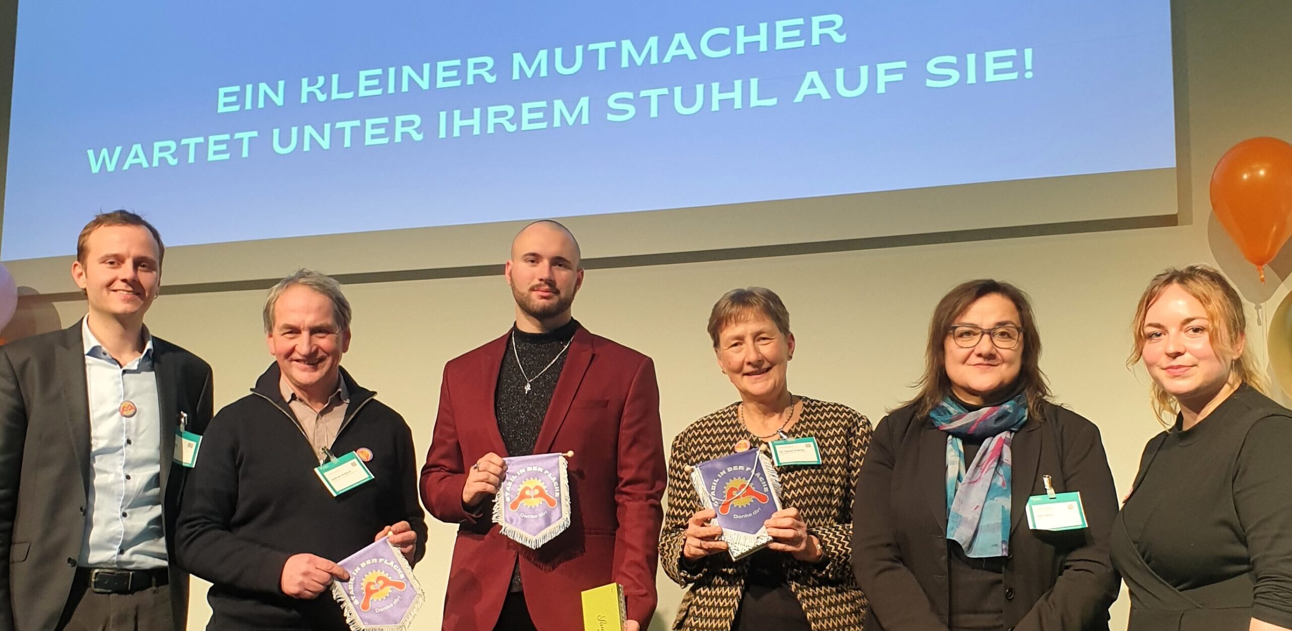 Sechs Personen stehen in einer Reihe auf einer Bühne. Die vier Personen in der Mitte halten lilafarbene Wimpel mit dem „Vielfalt:OST“-Logo und kleine Geschenke in den Händen – sie sind die Preisträger:innen.