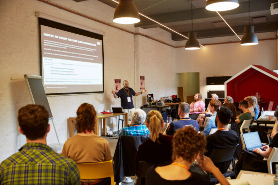 Eine männlich gelesene Person steht vorne links neben einer Projektionsfläche und spricht, mit beiden Armen gestikulierend, zu einer Gruppe von sitzenden Teilnehmenden. Alle sind sommerlich gekleidet, die meisten tragen orangefarbene Schlüsselbändern mit Namensschildern. Links steht ein leeres Flipchart, daneben ein Tisch mit Laptop und Materialien. Die Zuhörenden sitzen in mehreren Reihen, einige mit Laptops oder Notizblöcken. Im Hintergrund sind eine rote, hausförmige Nische und eine Küchenzeile erkennbar.