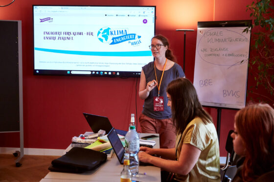 Eine weiblich gelesene Person steht vor einem Bildschirm mit der Webseite „Klima engagiert in Halle“ und spricht zu einer kleinen Gruppe. Neben ihr steht ein Flipchart mit der Aufschrift „Lokale Klimanetzwerke – wie Zugänge schaffen + ermöglichen?“. Zwei weitere Personen sitzen am Tisch und arbeiten an Laptops.