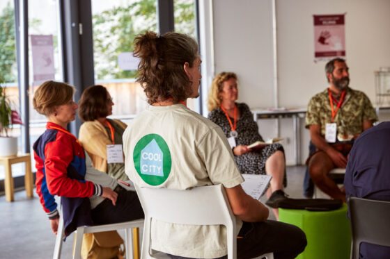 Mehrere Personen sitzen in einem Halbkreis auf Stühlen und hören zu, darunter eine Person im Vordergrund mit einem hellen T-Shirt mit der Aufschrift „COOL CITY“ auf dem Rücken. Sie tragen orangene Lanyards mit Namensschildern. Im Hintergrund sind große Fenster, Pflanzen und Poster an der weißen Wand zu sehen.