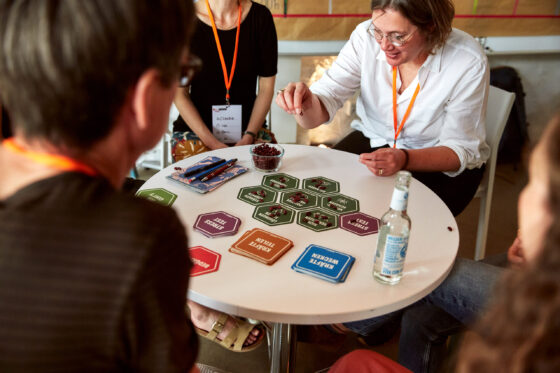 Mehrere Personen sitzen um einen runden Tisch und spielen ein Brettspiel mit farbigen Karten und sechseckigen Spielplättchen, auf denen kleine rote Bohnen liegen. Im Hintergrund sind ein Teil des Sessionplans und eine helle Wand erkennbar.