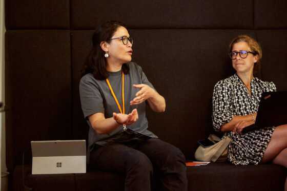 Zwei weiblich gelesene Personen sitzen auf einer schwarzen Polsterbank und sind in ein Gespräch vertieft. Die Person links gestikuliert lebhaft, während sie spricht. Die Person rechts hört aufmerksam zu, hält ein Laptop auf dem Schoß und wirkt nachdenklich.