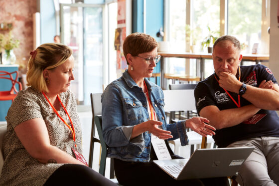 Drei Personen sitzen nebeneinander auf Stühlen um einen aufgeklappten Laptop. Die weiblich gelesene Person in der Mitte spricht und gestikuliert mit den Händen, während die beiden anderen aufmerksam zuhören. Alle tragen Namensschilder mit orangefarbenen Bändern um den Hals. Im Hintergrund sind große Fenster, durch die Tageslicht hereinfällt, sowie hohe Tische mit Pflanzen zu sehen.