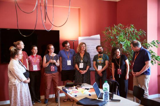Mehrere Personen stehen in einem Halbkreis in einem Raum mit roten Wänden und hören aufmerksam zu. Alle tragen Namensschilder an orangefarbenen Bändern, einige lächeln oder sind ins Gespräch vertieft. Im Vordergrund steht ein Tisch mit Laptops, Stiften und Notizzetteln, im Hintergrund ist ein Flipchart mit handgeschriebenen Notizen sichtbar.