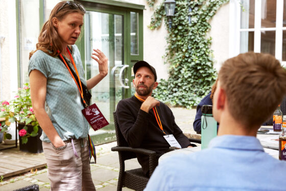 Eine weiblich gelesene Person steht und spricht zu einer kleinen Gruppe, während sie gestikuliert. Eine männlich gelesene Person mit schwarzem Oberteil und Kappe sitzt auf einem Stuhl und schaut aufmerksam zu ihr hoch. Im Vordergrund ist der Hinterkopf einer weiteren Person zu sehen, auf dem Tisch davor stehen Getränkeflaschen und eine Trinkflasche. Alle drei befindden sich in einem Außenbereich vor einem Gebäude mit Glastüren, Efeu an der Wand und Blumenkübeln.