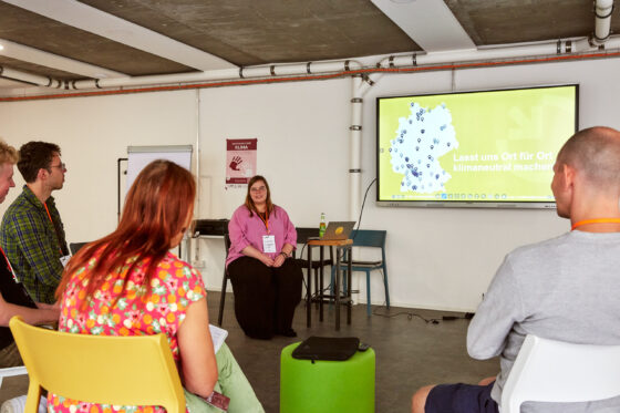 Eine weiblich gelesene Person im pinken Oberteil sitzt vorne in einem Stuhlkreis. Sie lächelt in die Runde, während im Hintergrund auf einem Bildschirm eine Deutschlandkarte mit Markierungen und der Satz „Lasst uns Ort für Ort klimaneutral machen“ eingeblendet ist. Vier weitere Personen sitzen im Halbkreis und hören aufmerksam zu, einige mit Notizblock in der Hand. Der Raum ist schlicht eingerichtet mit weißen Wänden und einigen Stühlen.