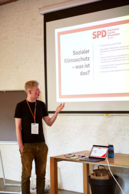 Eine männlich gelesene Person mit schwarzem T-Shirt und orangefarbenem Schlüsselband steht vor einer Leinwand und zeigt auf eine Präsentationsfolie mit der Überschrift „Sozialer Klimaschutz – was ist das?“. Im Hintergrund sind eine weiße Wand, ein Flipchart und ein Holztisch mit Laptop und Stiften zu sehen.