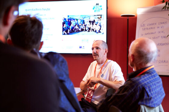 Eine männlich gelesene Person mit weißem T-Shirt und orangefarbenem Schlüsselband spricht in einer Sitzrunde; im Hintergrund sind ein Bildschirm mit der Überschrift „querstadtein heute“ und einem Gruppenfoto sowie ein Flipchart mit handschriftlichem Text zu sehen.