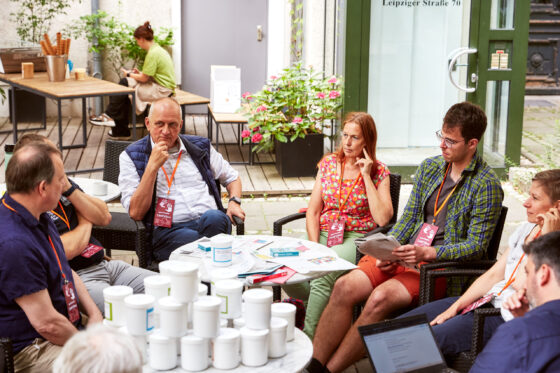 Mehrere Personen sitzen in einem Stuhlkreis um einen runden Tisch mit zahlreichen weißen Behältern und Papieren; alle tragen orangefarbene Schlüsselbänder mit der Aufschrift „openTransfer CAMP Klima“. Im Hintergrund sind Holztische und Pflanzen sowie eine sitzende Person in grünem Oberteil zu sehen.