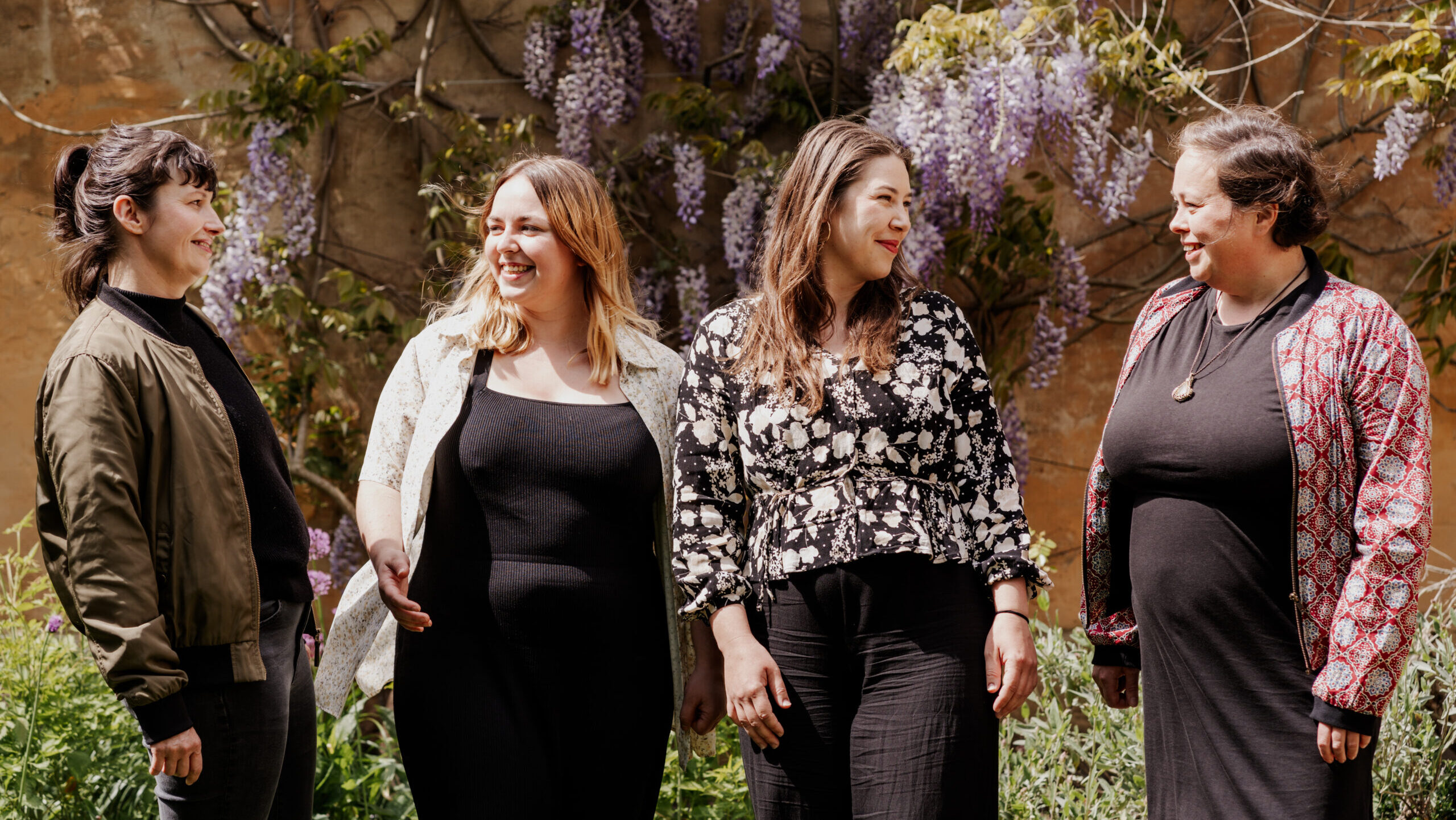 Teamfoto von Team openTransfer Zusammenhalt: Vier weiblich gelesene Personen stehen lachend in einem Garten vor einer mit blühendem Blauregen bewachsenen Wand. Sie blicken einander an und wirken fröhlich und verbunden.