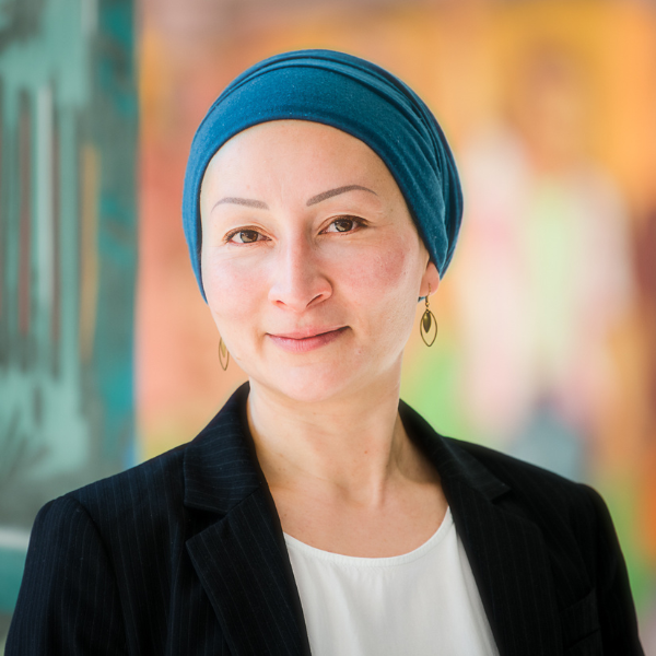 Porträt Sujin Noël: Eine weiblich gelesene Person mit blauem Kopftuch und schwarzem Blazer lächelt in die Kamera, der Hintergrund ist bunt und unscharf.