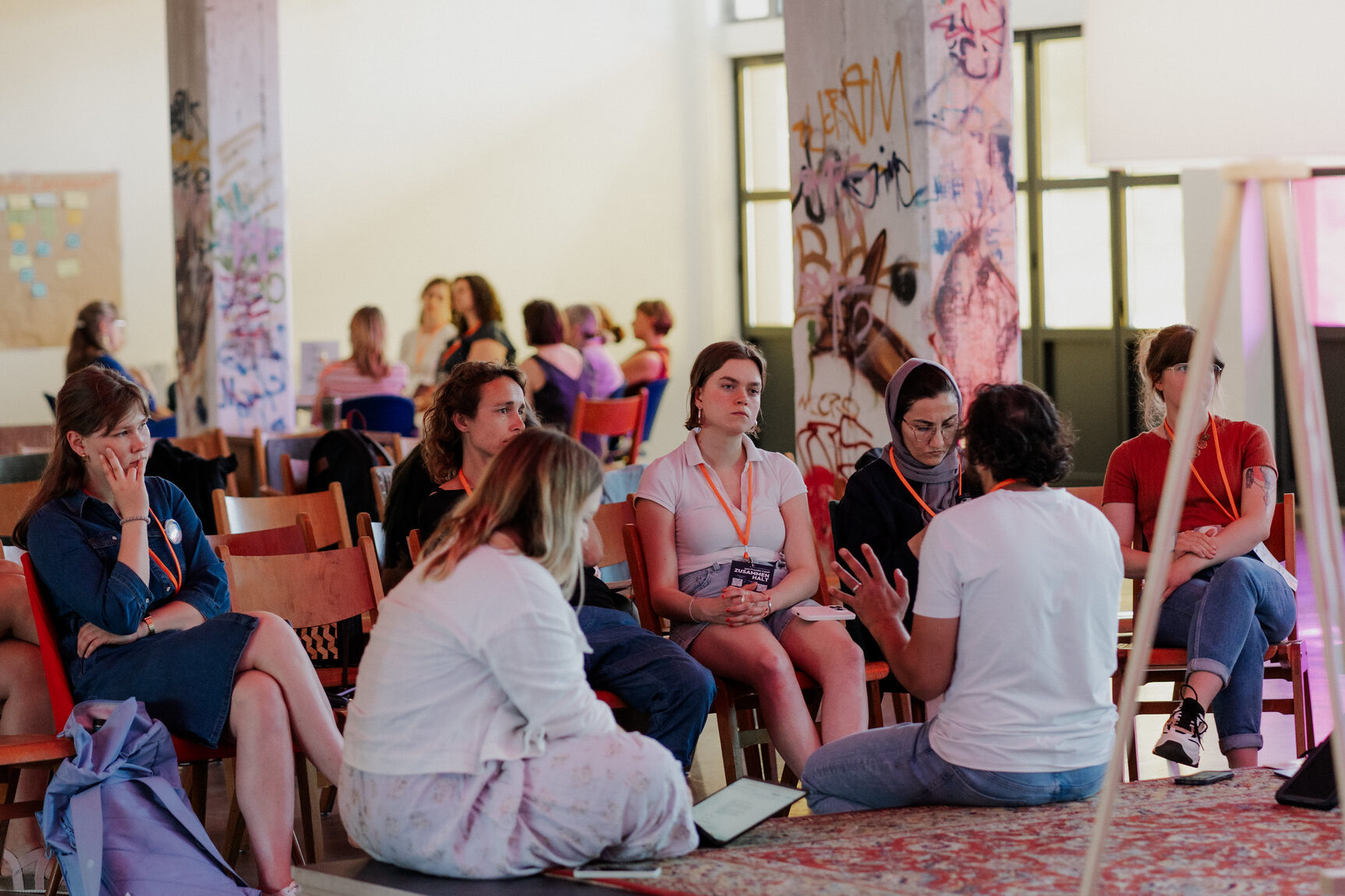 Mehrere Personen sitzen in einem Stuhlkreis in einem großen Raum mit bemalten Säulen; eine Person spricht, andere hören zu oder machen Notizen