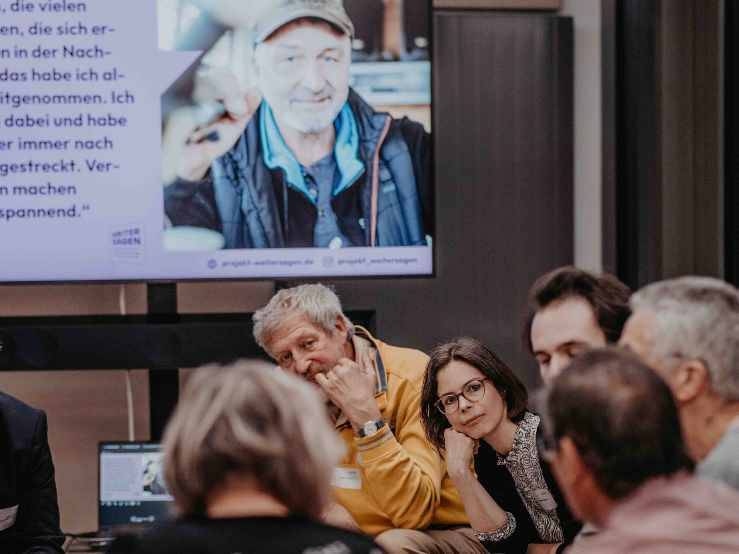 Mehrere Personen sitzen im Kreis und diskutieren. Im Hintergrund zeigt ein Bildschirm ein Foto einer älteren männlich gelesenen Person mit Mütze sowie einen längeren Text.